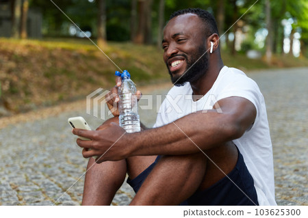 Young adult man resting on a park after a run with a bottle of water and smartphone. Outdoor workout Young adult man resting on a park after a run with a bottle of water and smartphone. Outdoor workout 103625300