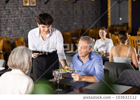 Young male waiter serving dish to elderly couple in restaurant 103632112