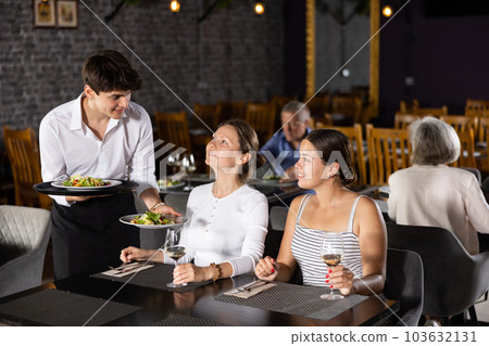 Two young women enjoying lunch in cozy restaurant, where waiter attentively serves them. Two young women enjoying lunch in cozy restaurant, where waiter attentively serves them. 103632131