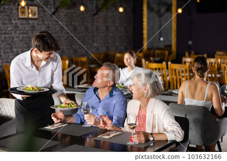 Young male waiter serving dish to elderly couple in restaurant 103632166