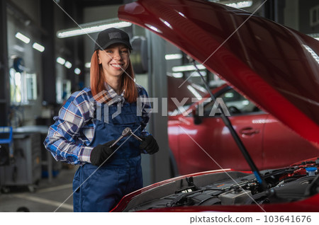 A woman auto mechanic stands at the open hood of a car and holds a screwdriver. A woman auto mechanic stands at the open hood of a car and holds a screwdriver. 103641676
