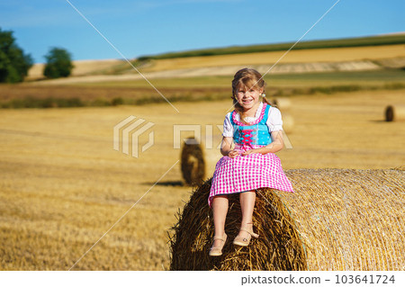 Cute little kid girl in traditional Bavarian costume in wheat field. Happy child with hay bale during Oktoberfest in Munich. Preschool girl play at hay bales during summer harvest time in Germany. Cute little kid girl in traditional Bavarian costume in wheat field. Happy child with hay bale during Oktoberfest in Munich. Preschool girl play at hay bales during summer harvest time in Germany. 103641724