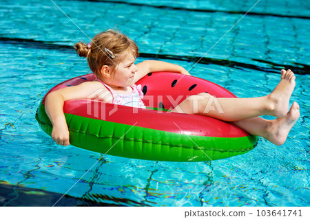 Happy little girl playing with colorful inflatable ring in outdoor swimming pool on sunny summer day. Preschool child learn to swim. Summer outdoor activity Happy little girl playing with colorful inflatable ring in outdoor swimming pool on sunny summer day. Preschool child learn to swim. Summer outdoor activity 103641741