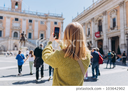 Blonde young Woman with smartphone is walking on a sunny day. Capitol in Rome, Piazza del Campidoglio in Capitoline Hill, Italy. Concept of traveling famous landmarks. 103642902