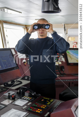 Officer on watch with binoculars on the navigational bridge. Caucasian man in blue uniform sweater on the bridge of cargo ship. 103643812