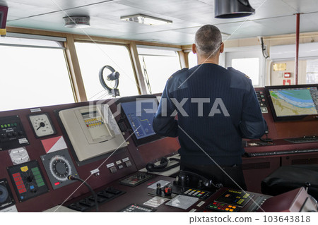 Officer on watch with binoculars on the navigational bridge. Caucasian man in blue uniform sweater on the bridge of cargo ship. 103643818