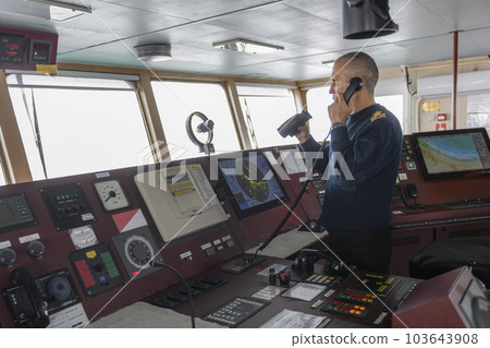 Officer on watch with radio on the navigational bridge. Caucasian man in blue uniform sweater on the bridge of cargo ship. Officer on watch with radio on the navigational bridge. Caucasian man in blue uniform sweater on the bridge of cargo ship. 103643908