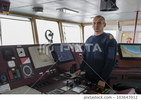 Officer on watch on the navigational bridge. Caucasian man in blue uniform sweater on the bridge of cargo ship. Officer on watch on the navigational bridge. Caucasian man in blue uniform sweater on the bridge of cargo ship. 103643912