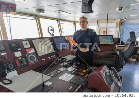 Officer on watch with SART on the navigational bridge. Caucasian man in blue uniform sweater using search and rescue radar transponder on the bridge of cargo ship. Officer on watch with SART on the navigational bridge. Caucasian man in blue uniform sweater using search and rescue radar transponder on the bridge of cargo ship. 103643925