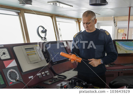 Officer on watch with SART on the navigational bridge. Caucasian man in blue uniform sweater using search and rescue radar transponder on the bridge of cargo ship. 103643929