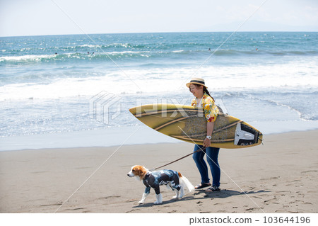 A woman with a surfboard and a dog wearing a rash guard 103644196