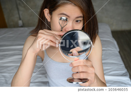 Portrait of young Asian woman using eyelash curler for curling eyelashes by looking mirror.  103646035