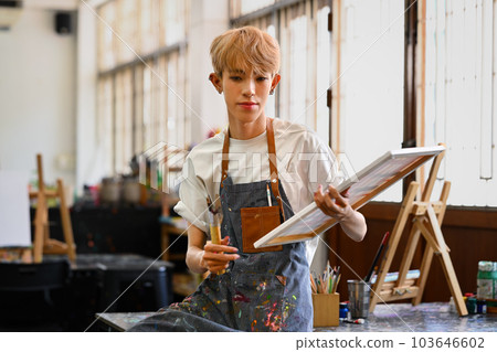Portrait of young man artist in apron holding art canvas and smiling to camera. Leisure activity, creative hobby and art concept 103646602