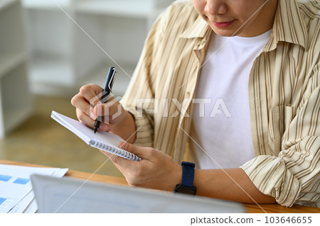 Young businessman looking at laptop screen and making important notes, planning daily appointment in diary Young businessman looking at laptop screen and making important notes, planning daily appointment in diary 103646655