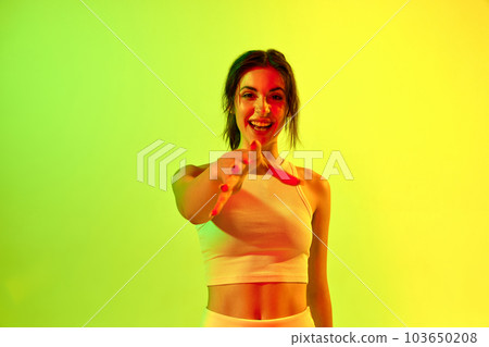 Image with positive brunette girl, young woman stretching hand to camera and smiling on acid green background in neon light. Happiness 103650208