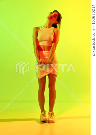 Portrait of happy, young woman with skinny body wearing elegant costume, skirt and top posing over pink background in neon light. Dreaming girl 103650214