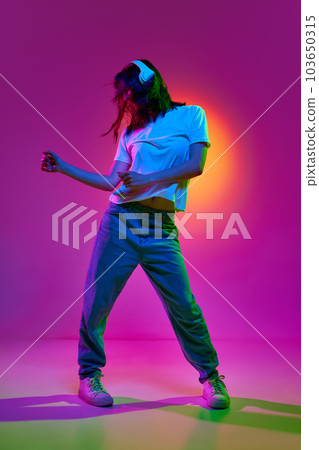 Portrait of young girl wearing casual clothes and headphones listening to music with joy and moving in beat of song over pink background in neon light 103650315