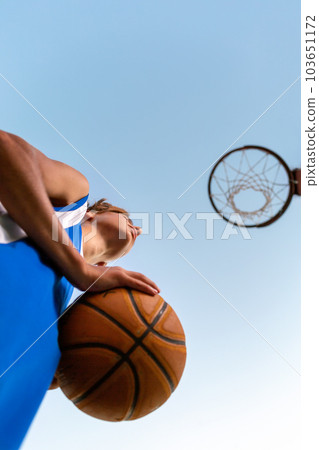 Basketball. A teenager in blue sportswear holds a ball in his hands and looks at a basketball Hoop. Bottom view. Sky in the background. Concept of sports games 103651172