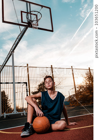 Sports and basketball. A young teenager in a black tracksuit poses with a basketball on a sports field while sitting on the ground. Bottom view Sports and basketball. A young teenager in a black tracksuit poses with a basketball on a sports field while sitting on the ground. Bottom view 103651200