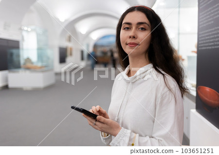 Portrait of young caucasian woman using smartphone in museum. Light hall in background. Concept of cultural exhibition and modern educational excursion Portrait of young caucasian woman using smartphone in museum. Light hall in background. Concept of cultural exhibition and modern educational excursion 103651251