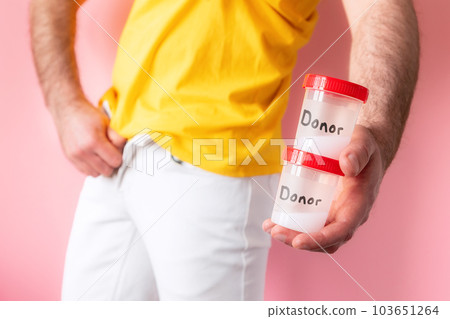 A man in white jeans zip up his fly and holds two plastic jars with sperm samples with text donor in his hands. Pink background. The concept of artificial insemination and sperm donation A man in white jeans zip up his fly and holds two plastic jars with sperm samples with text donor in his hands. Pink background. The concept of artificial insemination and sperm donation 103651264
