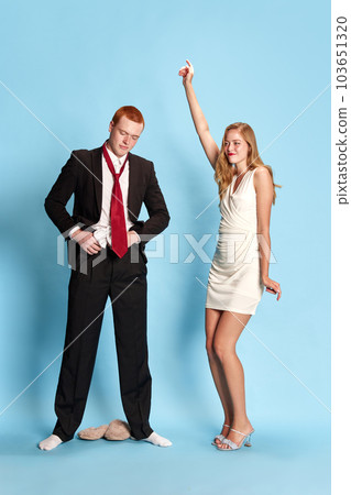 Portrait of beautiful young couple, man and woman getting ready to get married, woman dancing, man putting clothes on against blue studio background Portrait of beautiful young couple, man and woman getting ready to get married, woman dancing, man putting clothes on against blue studio background 103651320