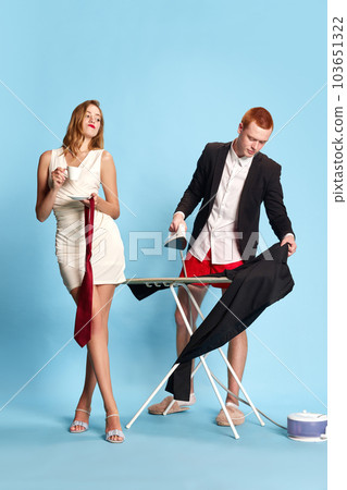 Getting ready. Pretty woman in white dress drinking coffee, young man in suit ironing his pants against blue studio background Getting ready. Pretty woman in white dress drinking coffee, young man in suit ironing his pants against blue studio background 103651322