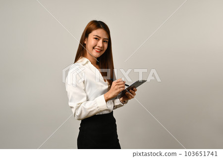 Charming young businesswoman holding digital tablet and smiling to camera isolated on gray background Charming young businesswoman holding digital tablet and smiling to camera isolated on gray background 103651741