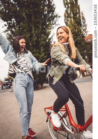 Vertical photo of two young women having fun in the city, one riding a red bike while other smiles with hands in the air behind her Vertical photo of two young women having fun in the city, one riding a red bike while other smiles with hands in the air behind her 103653438