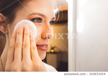 Close up side view of young woman removing make up off her face using a cotton pad and micellar water  103653454