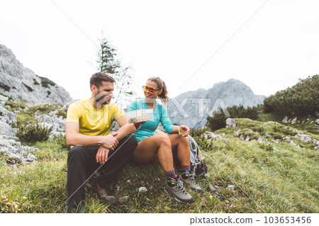 Couple of hikers sitting down on a grass field up in the mountains taking a stop for lunch break Couple of hikers sitting down on a grass field up in the mountains taking a stop for lunch break 103653456