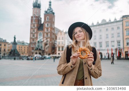 Attractive young female tourist with stylish clothes and a boho hat eating bagel obwarzanek traditional polish cuisine snack waling on Market square in Krakow. Traveling Europe Attractive young female tourist with stylish clothes and a boho hat eating bagel obwarzanek traditional polish cuisine snack waling on Market square in Krakow. Traveling Europe 103653498