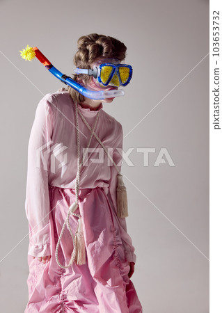 Portrait of young girl in pink outfit posing in water diving mask against grey studio background. Uniqueness and weirdness 103653732