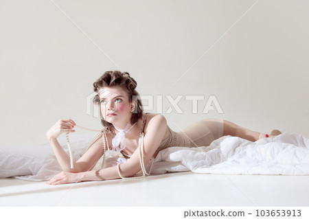 Beautiful, tender young girl with pink blusher on cheeks lying in retro lingerie on blanket against grey studio background. Sensuality 103653913