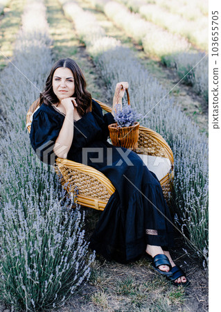 Attractive young woman in black dress sitting in chair surrounded by lavender field and holding basket with flowers 103655705