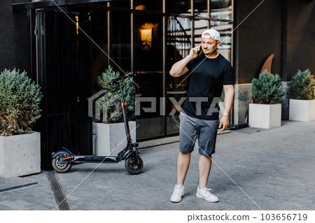 Caucasian young handsome man standing on street at electric scooter and talking on mobile phone. Stylish guy speaking on cellphone outdoors 103656719