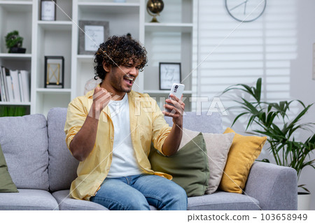Joyful and happy young man sitting on sofa alone at home, hispanic celebrating victory and success, got good news using app on phone. 103658949