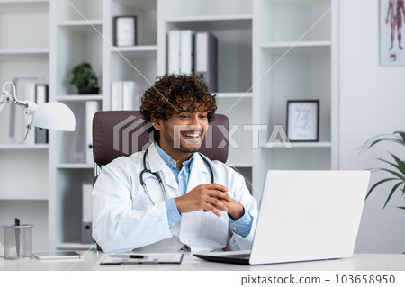 Young successful doctor working inside hospital office, man smiling and looking at laptop screen, hispanic man in medical gown is satisfied with patient's treatment achievement results. 103658950