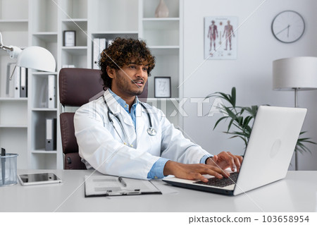 Young successful doctor working inside hospital office, man smiling and looking at laptop screen, hispanic man in medical gown is satisfied with patient's treatment achievement results. Young successful doctor working inside hospital office, man smiling and looking at laptop screen, hispanic man in medical gown is satisfied with patient's treatment achievement results. 103658954