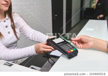 Close-up view of customer's hand paying by credit card and entering pin code on reader holded by a pleasant female manager administrator in the reception desk of modern medical clinic. NFC technology. 103660918