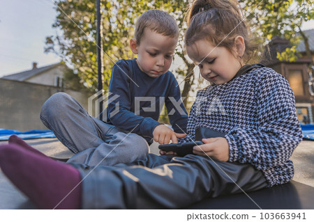 children sit on a trampoline and look at the phone children sit on a trampoline and look at the phone 103663941
