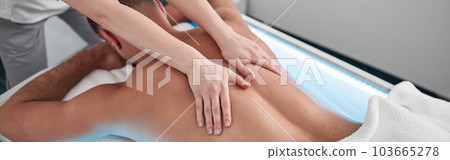 Specialist massages back of middle aged patient lying on glowing couch in clinic 103665278