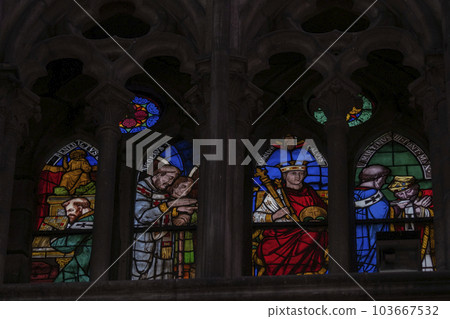 Multi colored medieval era stained glass panels depicting clergy, monarchs, saints set in stone gothic tracery in a cathedral. Illuminated windows against a dark background. Vivid colors 103667532