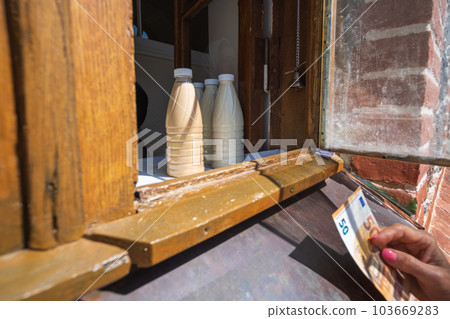 Country cow's milk in plastic bottles. A woman is holding a fifty euro banknote, buying fresh farm milk at an open window. ecologically clean product 103669283