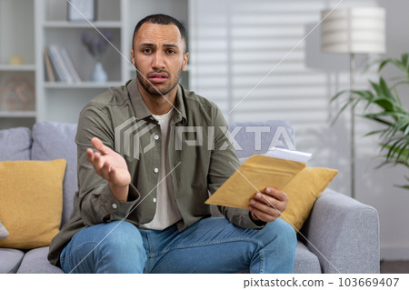 Portrait of an upset Latin American man who looks sadly into the camera and holds a letter with documents that he received in the mail with bad news. Worriedly spreading his hands. 103669407