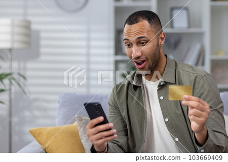 Shocked and happy young hispanic man using phone and credit card at home on sofa. Made a successful transaction, received a cash reward, online shopping. 103669409