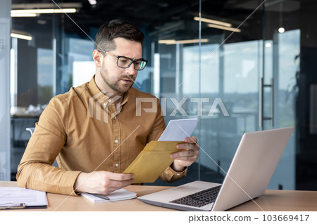 A serious and worried young businessman working in the office holds a letter, documents. got bad news. Saddenedly reads a message, a contract, an agreement. A serious and worried young businessman working in the office holds a letter, documents. got bad news. Saddenedly reads a message, a contract, an agreement. 103669417