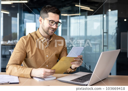 A young man businessman sits smiling in the office at the table and holds in his hands and reads documents, a contract, a successful deal that he received by mail. A young man businessman sits smiling in the office at the table and holds in his hands and reads documents, a contract, a successful deal that he received by mail. 103669418