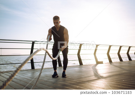 Young athlete wearing sports clothes is doing workout at the beach pier. Man doing exercises outdoors. 103671183