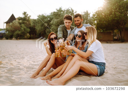 Group of friends cheers and drink beers on the beach. Young friends relaxing and having picnic. Suummertime. 103672298